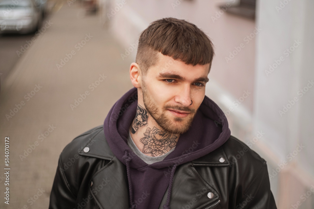Fototapeta premium Street portrait of a handsome stylish hipster guy with a hair and beard with a tattoo on his neck in fashionable clothes with a jacket and hoodie walking in the city