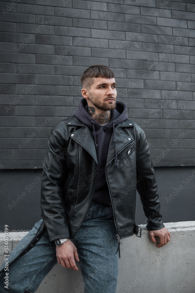 Fashionable handsome man with hairstyle and beard in stylish casual clothes with jacket and hoodie near a black brick wall in the city