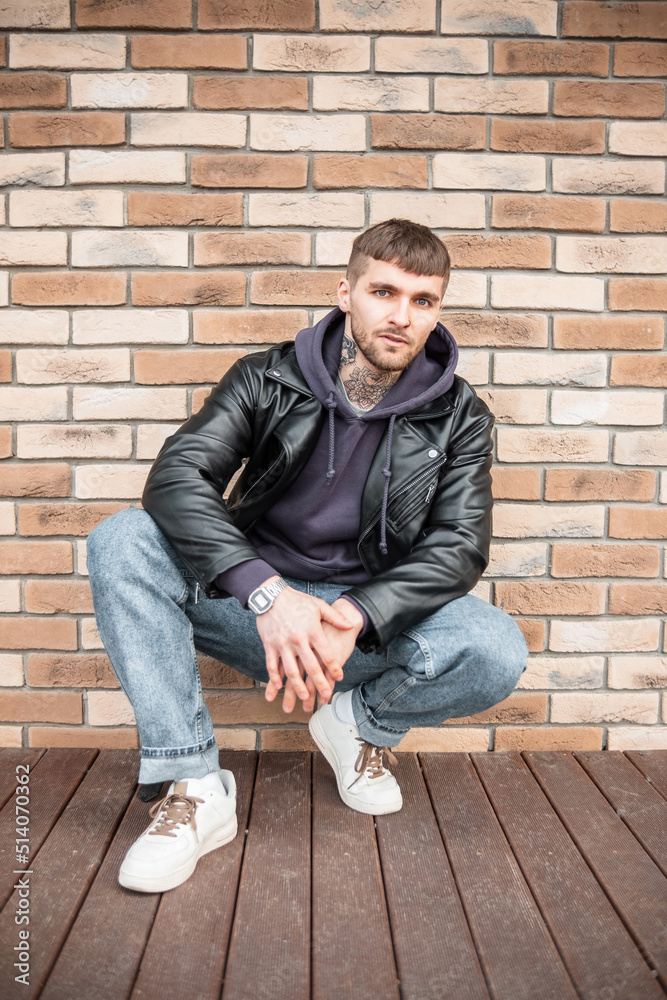 Cool trendy hipster guy in fashion casual clothes with leather jacket, jeans, hoodie and sneakers sits near a brick wall on the street