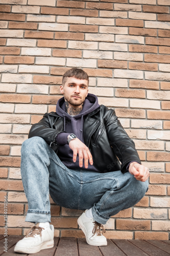 Cool handsome young stylish hipster man in fashionable clothes with leather jacket, hoodie, jeans and sneakers sitting outside near a brick wall Cool handsome young stylish hipster man in fashionable clothes with leather jacket, hoodie, jeans and sneakers sitting outside near a brick wall