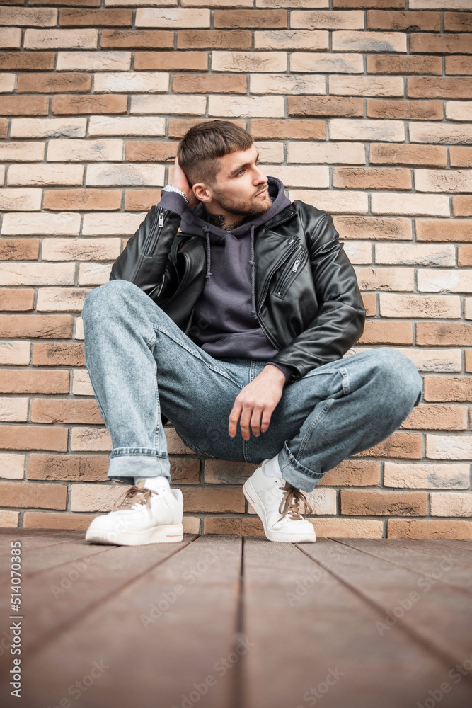 Fashionable handsome young man hipster model in a stylish outfit with leather jacket, hoodie, jeans and white sneakers sitting near a brick wall Fashionable handsome young man hipster model in a stylish outfit with leather jacket, hoodie, jeans and white sneakers sitting near a brick wall