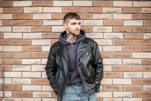 Fashionable hipster man with a tattoo on his neck in stylish casual clothes with a jacket and hoodie standing near a brick wall on the street