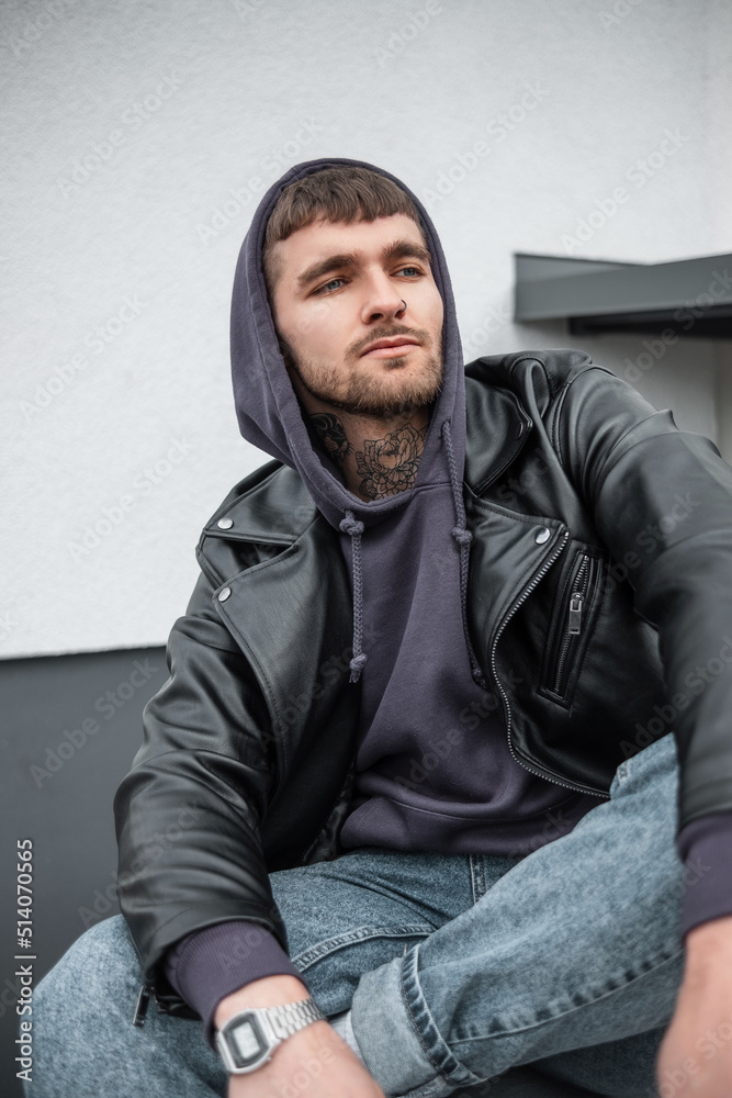 Handsome trendy hipster model guy with hair and a beard in a fashionable leather rock jacket with hoodies and jeans sits and rest near a white building on the street Handsome trendy hipster model guy with hair and a beard in a fashionable leather rock jacket with hoodies and jeans sits and rest near a white building on the street