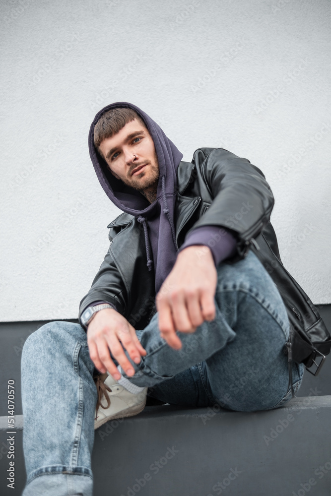 Cool fashion trendy handsome man model with rock fashion leather jacket, hoodie and jeans sits near a white wall on the street
