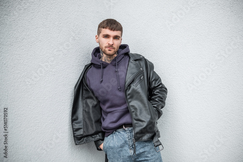 Fashionable street handsome hipster man in fashionable casual clothes with a leather jacket and hoodie stands near a white wall on the street. © alones