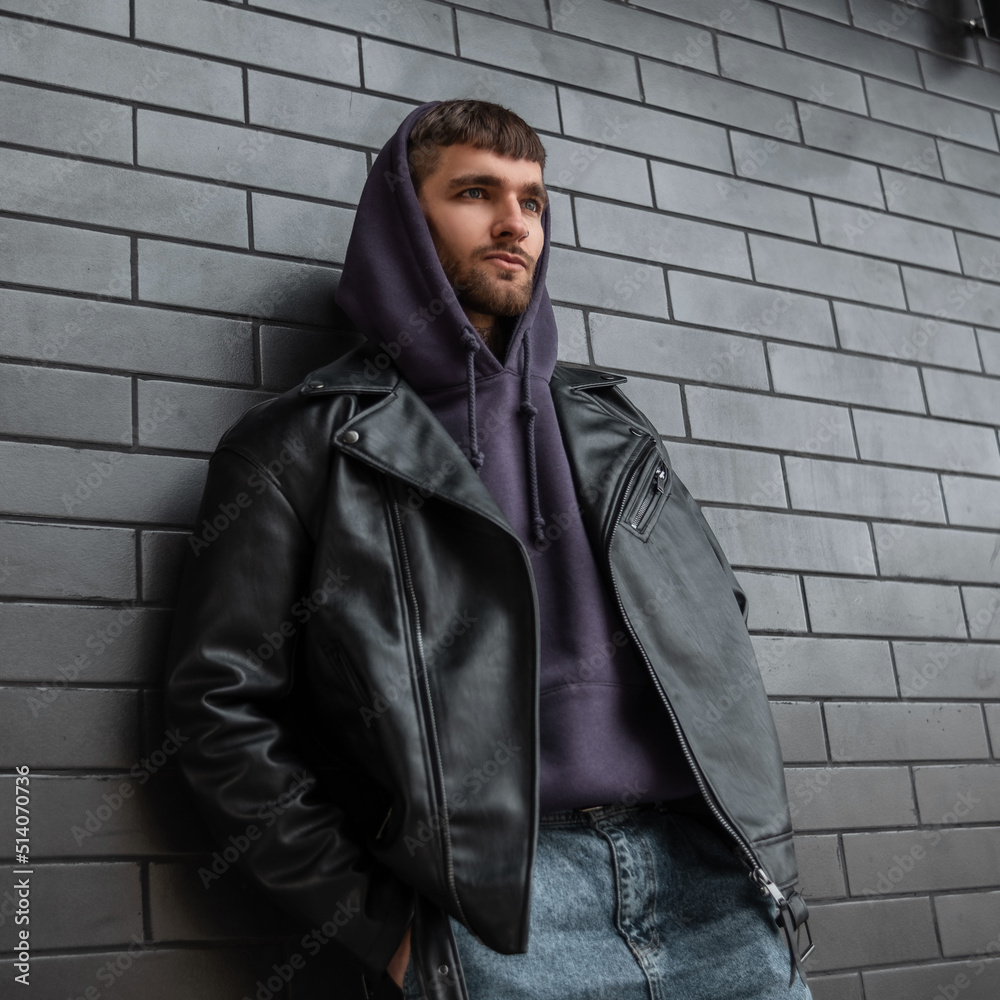 Stylish handsome brutal guy in fashionable casual clothes with a leather rock jacket and hoodie standing near a black brick wall on the street Stylish handsome brutal guy in fashionable casual clothes with a leather rock jacket and hoodie standing near a black brick wall on the street