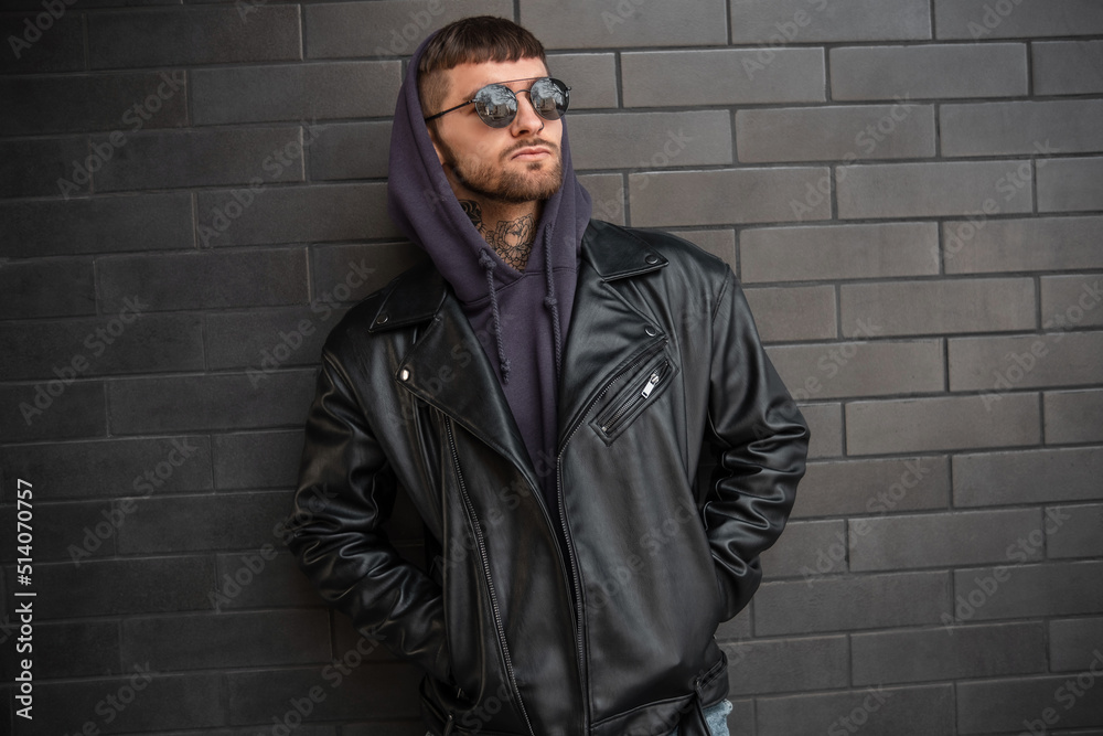 Fashion street handsome man model hipster with round vintage sunglasses in black fashionable urban outfit with rock leather jacket and hoodie stands near a black brick wall Fashion street handsome man model hipster with round vintage sunglasses in black fashionable urban outfit with rock leather jacket and hoodie stands near a black brick wall