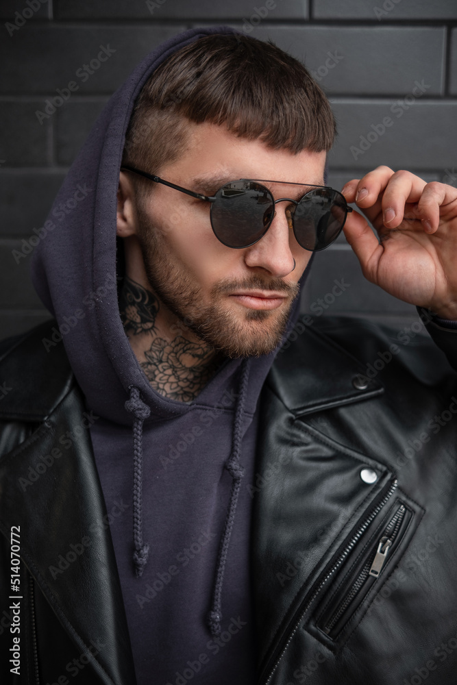 Obraz premium Fashion street male portrait of a handsome business stylish man in fashionable black sunglasses with a hood in a trendy black leather jacket and hoodie on a dark brick background on the street