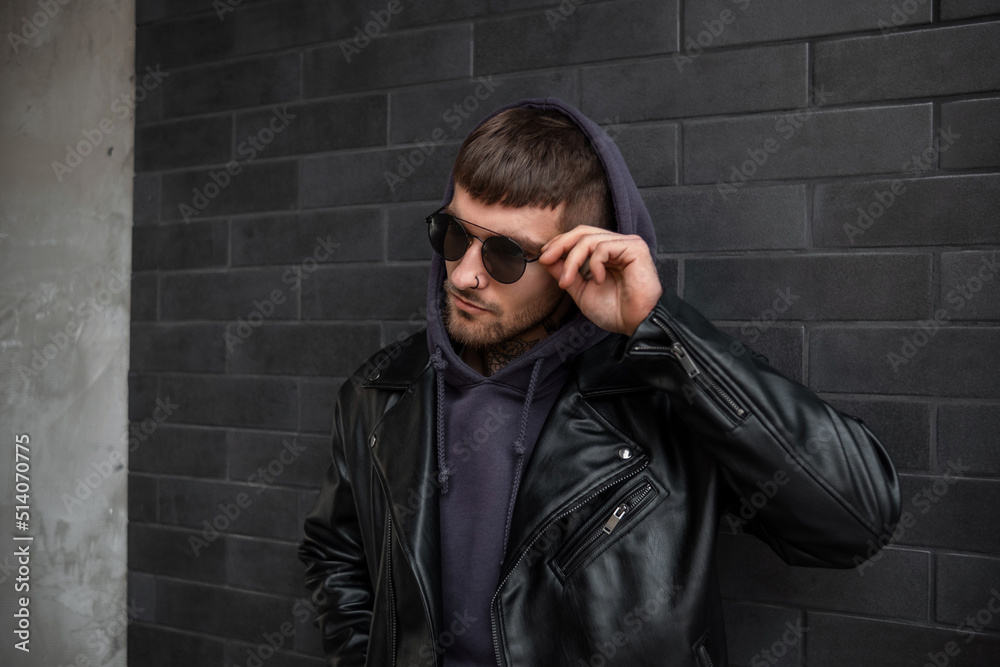 Fashionable handsome guy in stylish black clothes with a leather jacket and hoodie is fixing his sunglasses and standing near a black brick wall on the street