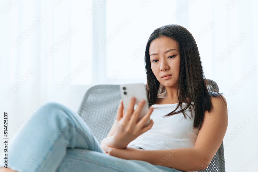 Upset confused tanned lovely young Asian woman read shocked message using phone at home interior living room. Distance communication, Remote work, Social media concept. Cool offer Banner Wide angle Upset confused tanned lovely young Asian woman read shocked message using phone at home interior living room. Distance communication, Remote work, Social media concept. Cool offer Banner Wide angle