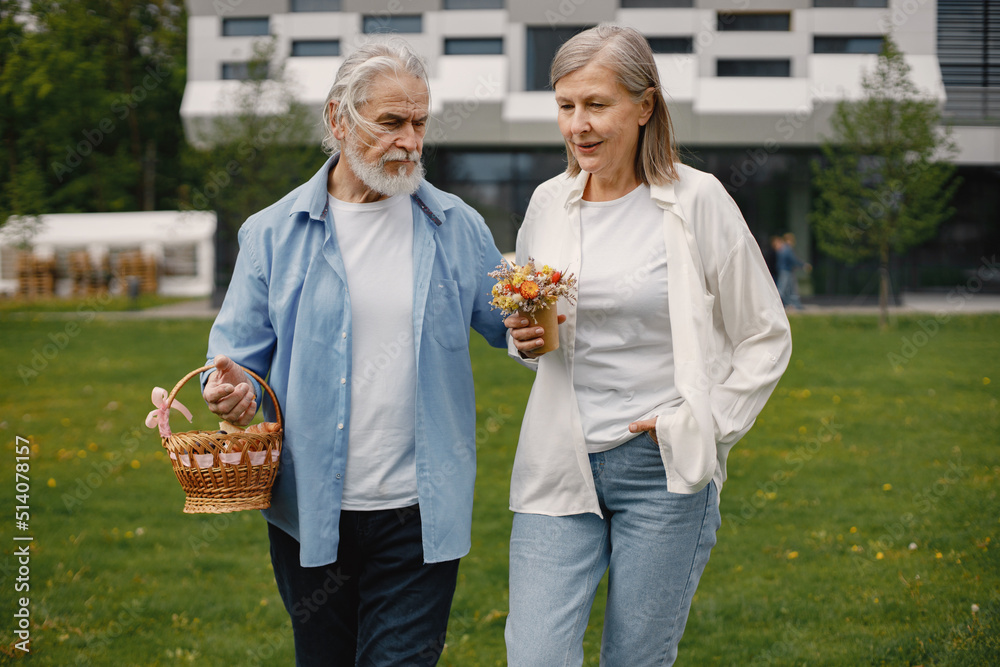 Senior couple standing on a grass in summer with straw basket and flowers Senior couple standing on a grass in summer with straw basket and flowers