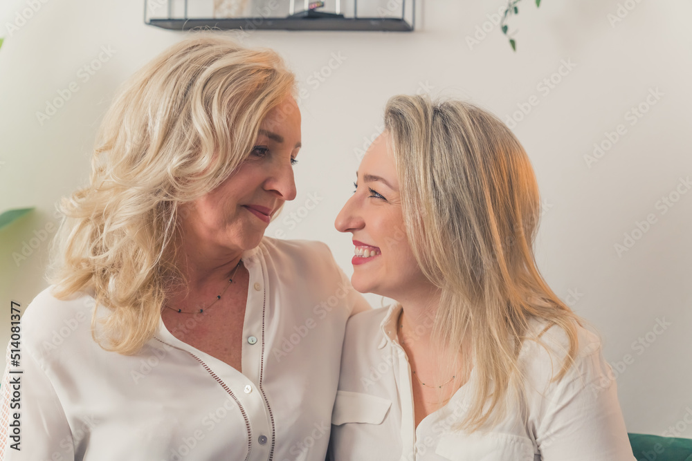 Indoor shot in an apartment of two joyful caucasian blonde-haired women sitting on a couch looking at each other and smiling, waering white chic shirts. High quality photo Indoor shot in an apartment of two joyful caucasian blonde-haired women sitting on a couch looking at each other and smiling, waering white chic shirts. High quality photo