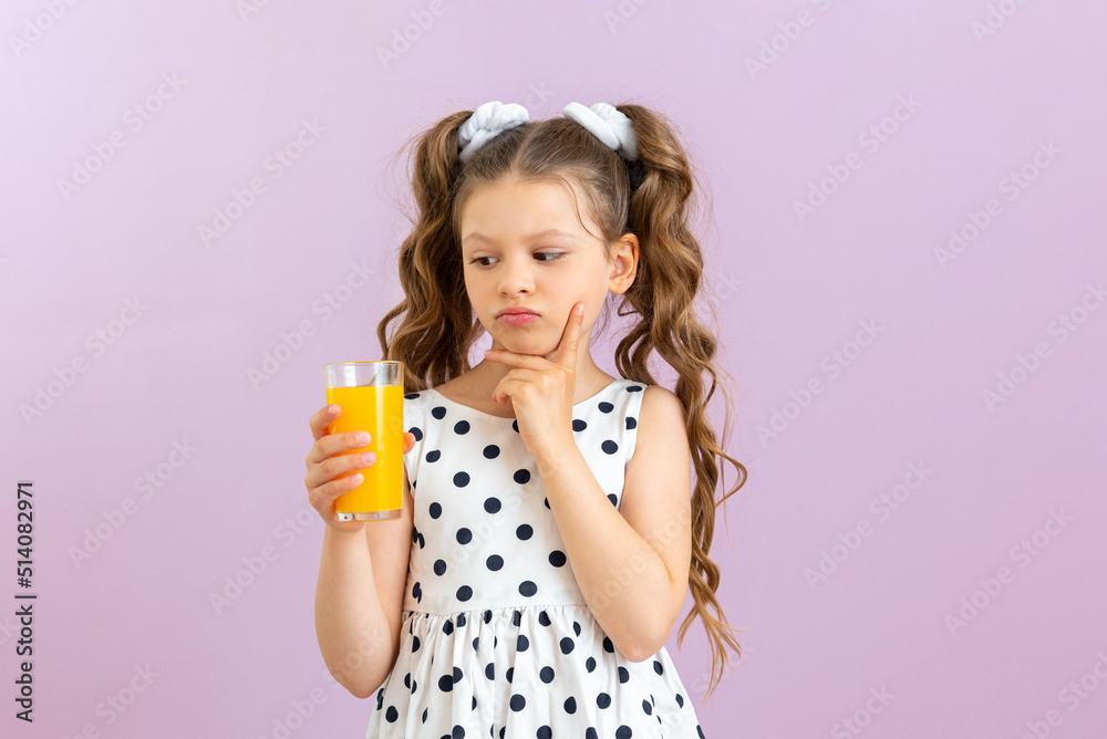 A glass of orange juice in the child's hands. A little girl in a polka dot dress holds citrus juice on a pink isolated background. Vitamins for children. A glass of orange juice in the child's hands. A little girl in a polka dot dress holds citrus juice on a pink isolated background. Vitamins for children.