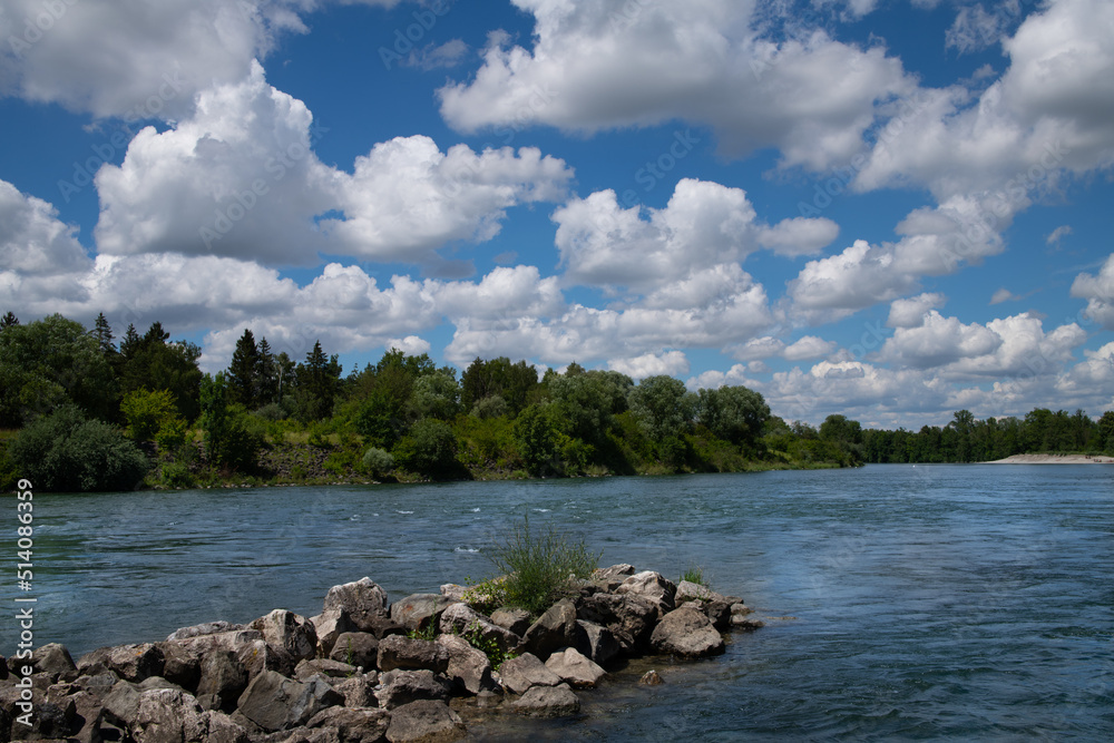 Wide scenic shot of a large river that flows in Germany, the Lech. In the blue sky are many white clouds. At the edge of the water are many trees.