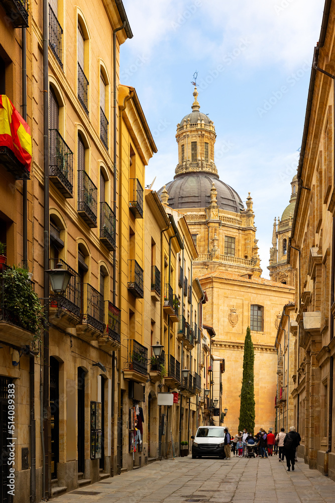 Picturesque cityscape of historic area of Salamanca overlooking people walking along typical narrow paved street leading to baroque building of La Clerecia with huge dome on sunny spring day, Spain.