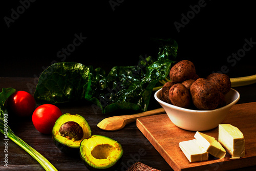 Rustic potatoes, sliced cheese on a wooden board, next to a sliced avocado, tomatoes and Swiss chard