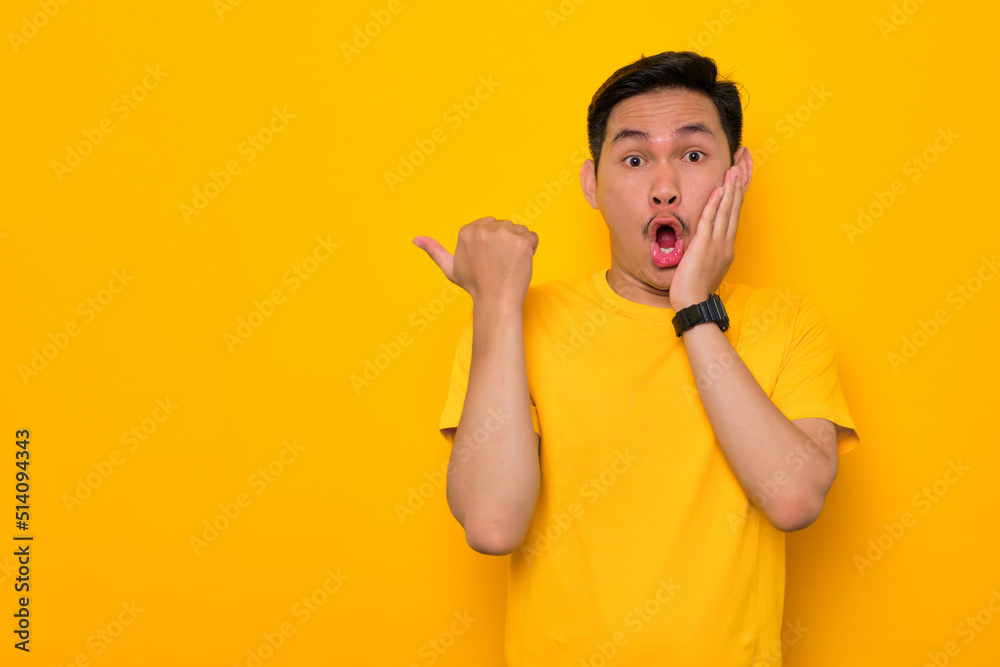 Shocked young Asian man in casual t-shirt pointing thumb aside at copy space isolated on yellow background. Great offer concept Shocked young Asian man in casual t-shirt pointing thumb aside at copy space isolated on yellow background. Great offer concept