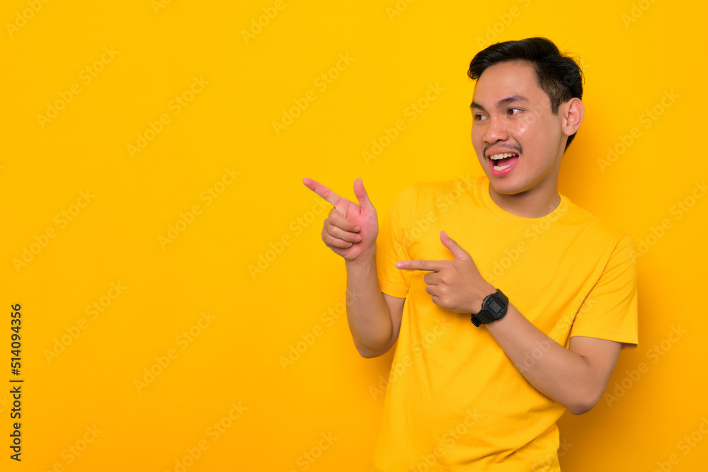 Excited young Asian man in casual t-shirt pointing finger aside at copy space isolated on yellow background. Great offer concept