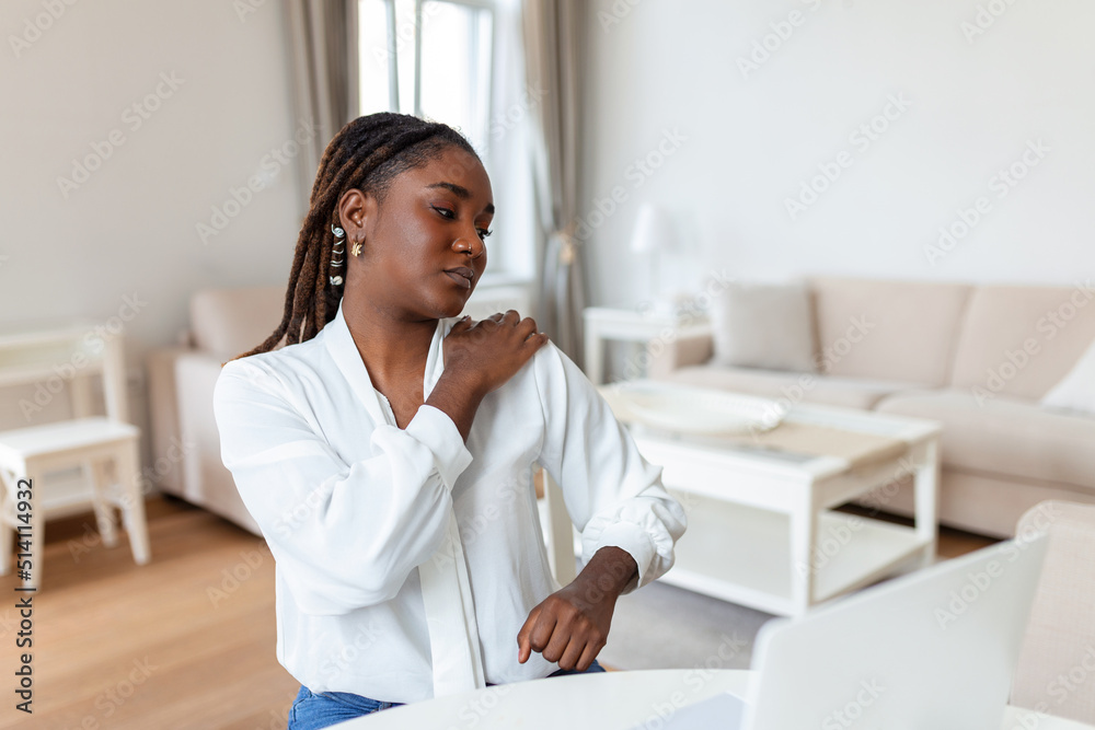 Portrait of young stressed African woman sitting at home office desk in front of laptop, touching aching shoulder with pained expression, suffering from shoulder ache after working on pc Portrait of young stressed African woman sitting at home office desk in front of laptop, touching aching shoulder with pained expression, suffering from shoulder ache after working on pc