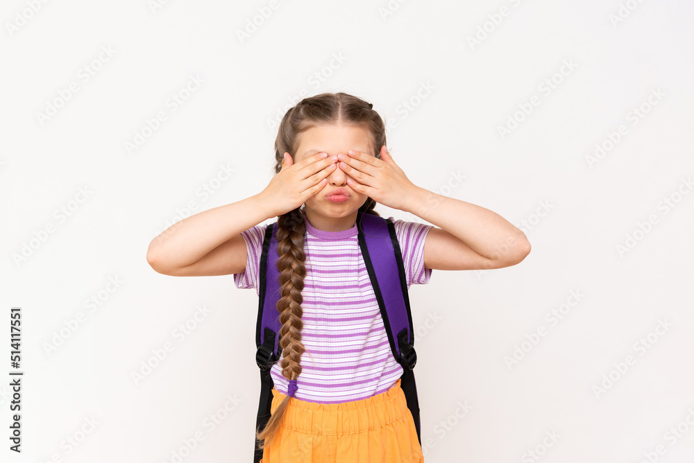 A little girl with a backpack on her back covers her eyes with her hands and smiles broadly on a white isolated background. School education for children. A little girl with a backpack on her back covers her eyes with her hands and smiles broadly on a white isolated background. School education for children.