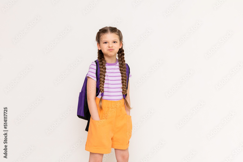 A little girl with a backpack on her back puffed out her cheeks and put her hands in the pockets of her shorts on a white isolated background. A little girl with a backpack on her back puffed out her cheeks and put her hands in the pockets of her shorts on a white isolated background.