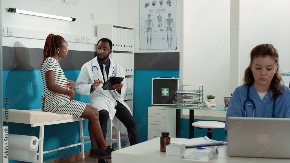 Obraz premium Obstetrician using digital tablet to consult patient expecting child at medical appointment. Male doctor taking notes at checkup visit examination, talking to woman with baby bump.