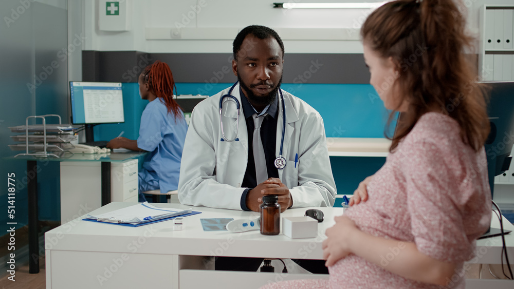 Obraz premium Male obstetrician having checkup appointment with pregnant patient, talking about maternity and childbirth in medical cabinet. Health specialist consulting woman with pregnancy belly.