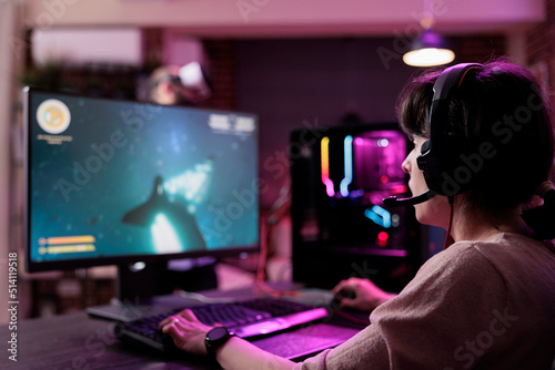 Modern person playing video games online on live stream, using computer and neon lights in living room. Female gamer having fun with action gameplay on virtual shooting tournament.