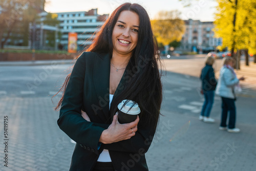 Happy 30s beautiful caucasian business woman drinking hot coffee holding paper cup while walking in city sunset park.