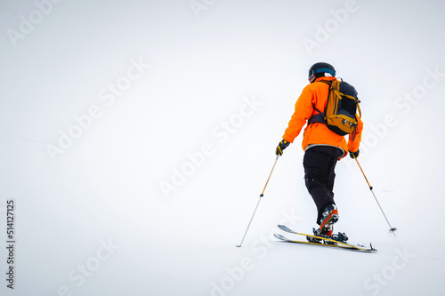 A male athlete in an orange jacket with a backpack on his shoulders and a helmet with a mask in a ski tour climbs a snowy slope in foggy weather. Ski touring and skiing copy space