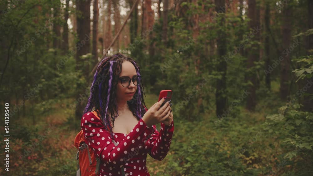 Traveling woman with dreadlocks with a phone in woods. Female traveler with backpack in woods reading map on a smartphone and looking for a way in cloudy weather Traveling woman with dreadlocks with a phone in woods. Female traveler with backpack in woods reading map on a smartphone and looking for a way in cloudy weather