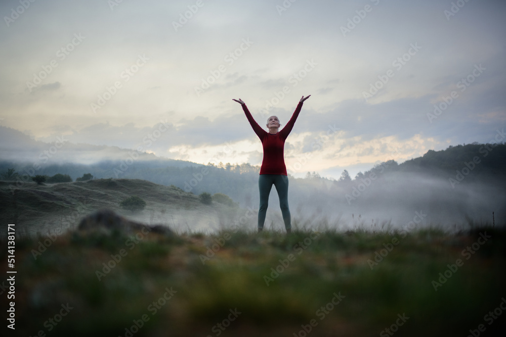 Senior woman doing breathing exercise in nature on early morning with fog and mountains in background.