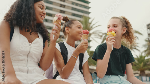 Laughing three girls friends pre-teenage are sitting on the waterfront testing ice cream. Three teenagers enjoy ice cream on hot summer day.