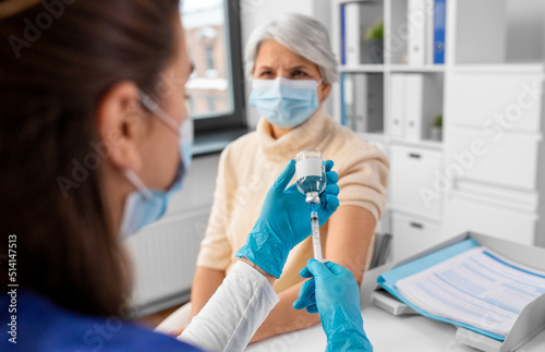 medicine, health and vaccination concept - close up of doctor or nurse filling syringe with vaccine or drug and senior woman in mask at hospital