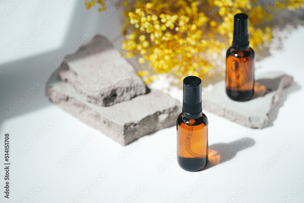 Amber pump bottles with serum, tonic or essential oil on grey concrete podium with yellow flowers. White background with daylight. Beauty concept for face and body care Amber pump bottles with serum, tonic or essential oil on grey concrete podium with yellow flowers. White background with daylight. Beauty concept for face and body care