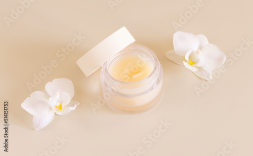 Cosmetic glass jar near white orchid flowers on light yellow close up