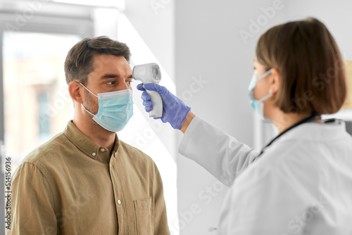 medicine, healthcare and people concept - female doctor in mask with infrared forehead thermometer measuring temperature of man patient at hospital