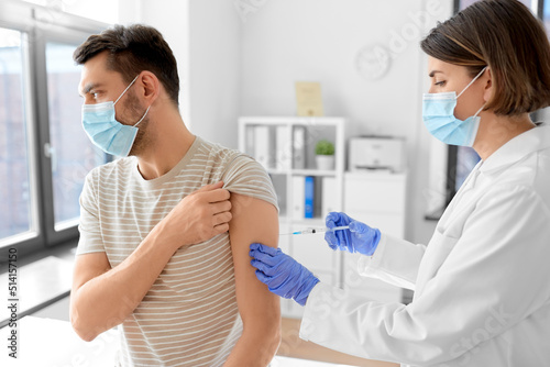 health, medicine and pandemic concept - female doctor or nurse wearing protective medical mask and gloves with syringe vaccinating male patient at hospital