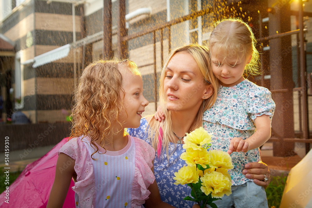 Two cute little blonde girls sisters and mother with flowers on a green lawn under rain drops in a summer sunny day near rural house in a village. Happy family having rest and fun in good spring day Two cute little blonde girls sisters and mother with flowers on a green lawn under rain drops in a summer sunny day near rural house in a village. Happy family having rest and fun in good spring day