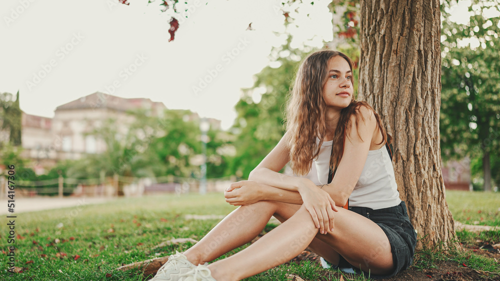 Beautiful girl with long wavy hair in white top looks around while sitting on the green grass under tree in the park