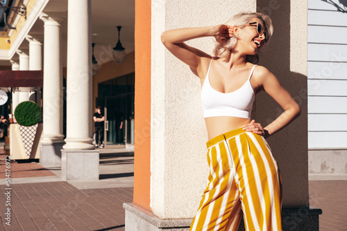 Young beautiful smiling hipster woman in trendy summer clothes. Sexy carefree model posing on the street background at sunset. Positive blond female. Cheerful and happy. In sunglasses