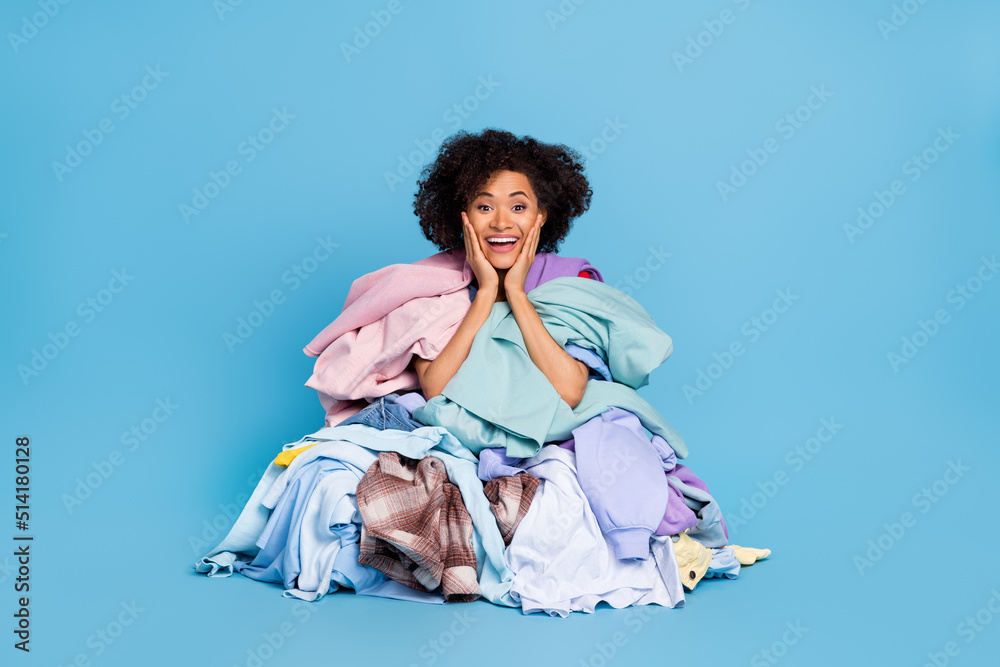 Photo of positive excited woman arms hands cheekbones cheeks covering big clothes stack isolated blue color background Photo of positive excited woman arms hands cheekbones cheeks covering big clothes stack isolated blue color background