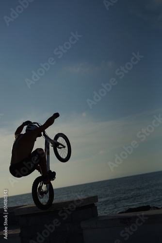 Trial biker performing on the rocks. Experienced biker does agility tricks on the beach in front of the setting sun. Shirtless man and trial bunny hops on the sea.
