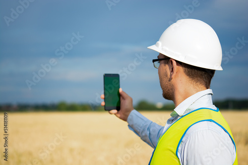 an engineer is working on his smartphone in the field, an agricultural engineer is standing in the field, an engineer is holding a phone in his hand, an engineer is wearing a helmet