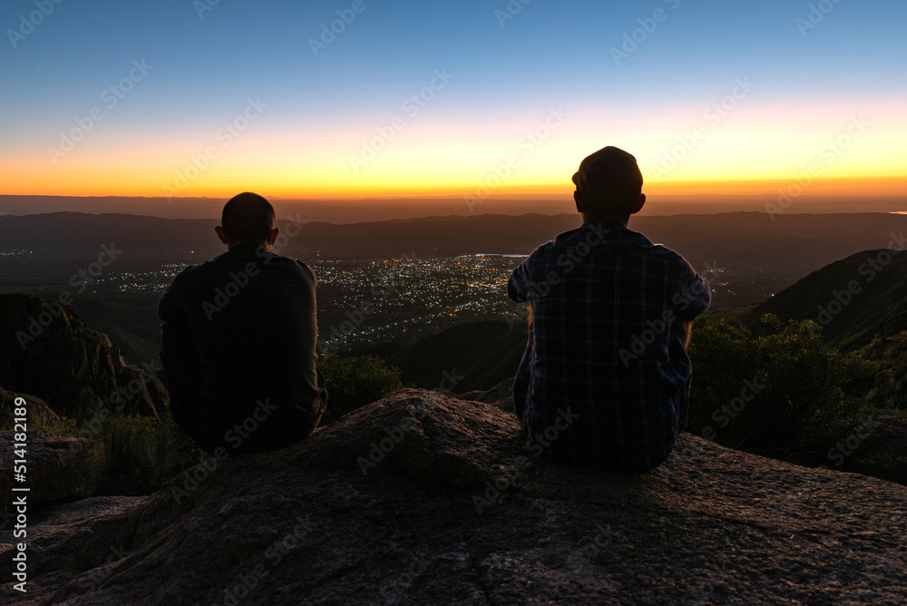 Rear View of Two Young Man Sillhouette Sitting on a Cliff Watching the Sunset from a Mountain