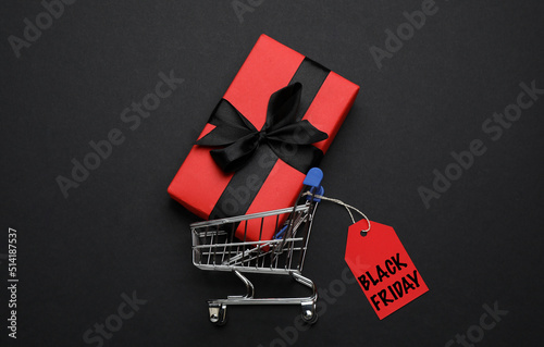Shopping cart, tag with phrase BLACK FRIDAY and gift box on dark background, flat lay
