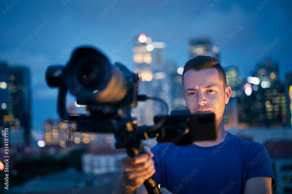 Man filming with camera and gimbal. Portrait of videographer against illuminated urban skyline at night.. Man filming with camera and gimbal. Portrait of videographer against illuminated urban skyline at night..