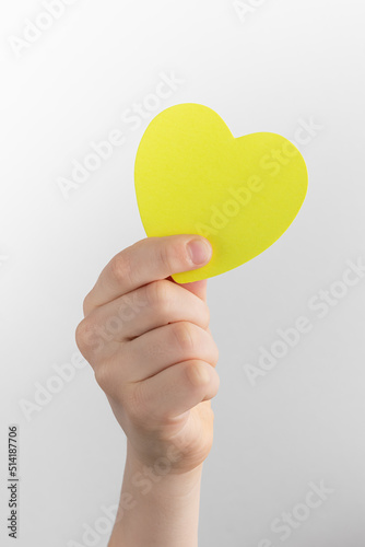 child hand holding yellow blank reminder or paper notes in the shape of a heart above a white and gray background, copy space