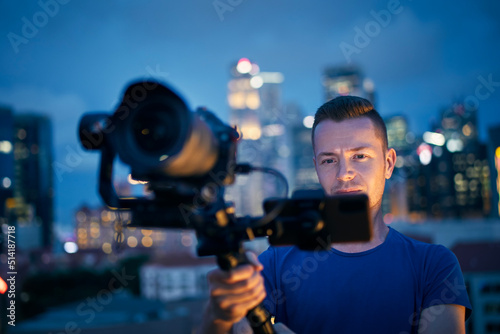 Man filming with camera and gimbal. Portrait of videographer against illuminated urban skyline at night..