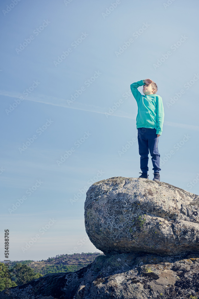 Preteen boy having fun looking to the horizon on the top of a huge rock while a journey day enjoying the nature hiking. Discover concept.